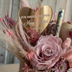 May include: A pink and gold heart-shaped sign with the text "F&uuml;r immer und ewig Margo & Robert 12.09.2025" surrounded by dried flowers in a pink container.