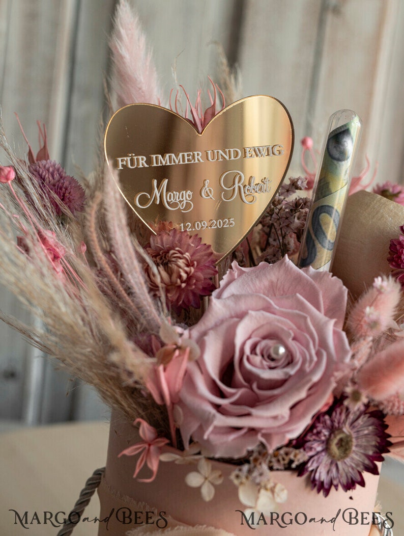 May include: A pink and gold heart-shaped sign with the text "F&uuml;r immer und ewig Margo & Robert 12.09.2025" surrounded by dried flowers in a pink container.