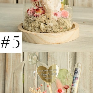 May include: A glass dome with a wooden base contains a preserved flower arrangement. The arrangement includes a white rose, pink dried flowers, and greenery. A gold heart-shaped plaque with the text "F&Uuml;R IMMER UND EWIG Margo & Robert 06.06.2026" is attached to a stem in the arrangement.