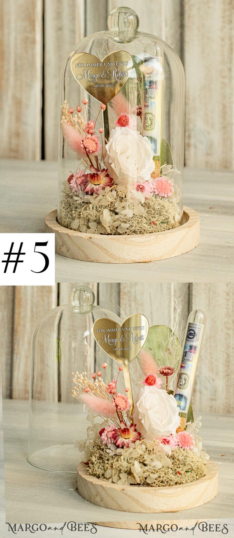 May include: A glass dome with a wooden base contains a preserved flower arrangement. The arrangement includes a white rose, pink dried flowers, and greenery. A gold heart-shaped plaque with the text "F&Uuml;R IMMER UND EWIG Margo & Robert 06.06.2026" is attached to a stem in the arrangement.