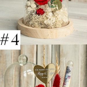 May include: A glass dome with a wooden base contains a preserved red rose, white and red dried flowers, and a gold heart-shaped plaque with the text "F&Uuml;R IMMER UND EWIG Margo & Robert 06.06.2026".
