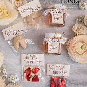 May include: A selection of wedding favors displayed on a wooden surface. The favors include fortune cookies, honey jars, and chocolate bars. The fortune cookies are wrapped in clear plastic bags with a tag that says "Gl&uuml;ck's Kek's". The honey jars are decorated with lace and a tag that says "Liebe Macht Das Leben S&uuml;per". The chocolate bars are wrapped in clear plastic bags with a tag that says "Liebe Macht Das Leben S&uuml;per".
