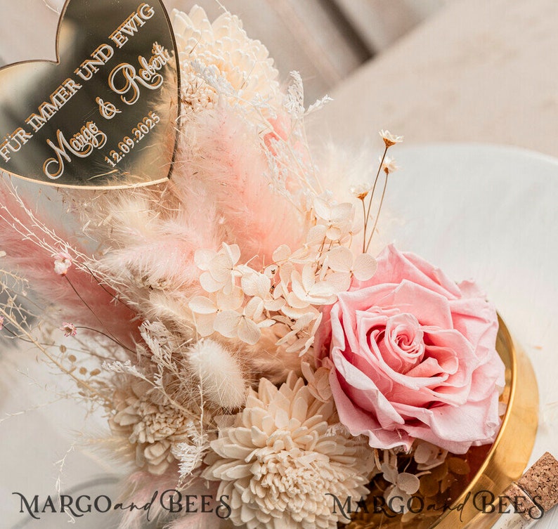 May include: A gold heart-shaped plaque with the text "F&uuml;r immer und ewig" and "Maig & Robert" with the date "12.09.2025" is placed on top of a bouquet of dried flowers. The bouquet includes pink and white dried flowers, a single pink rose, and dried pampas grass.