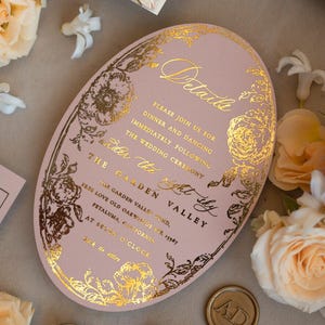 May include: A pink oval wedding invitation with gold foil detailing. The invitation features a floral design and the text "Details", "Please join us for dinner and dancing immediately following the wedding ceremony", "Under the right sky", "The Garden Valley", "The Garden Valley Fund, 15987 Love Old Oakwards St. Petaluma, California", "At Seven O'Clock", "Black tie attire".