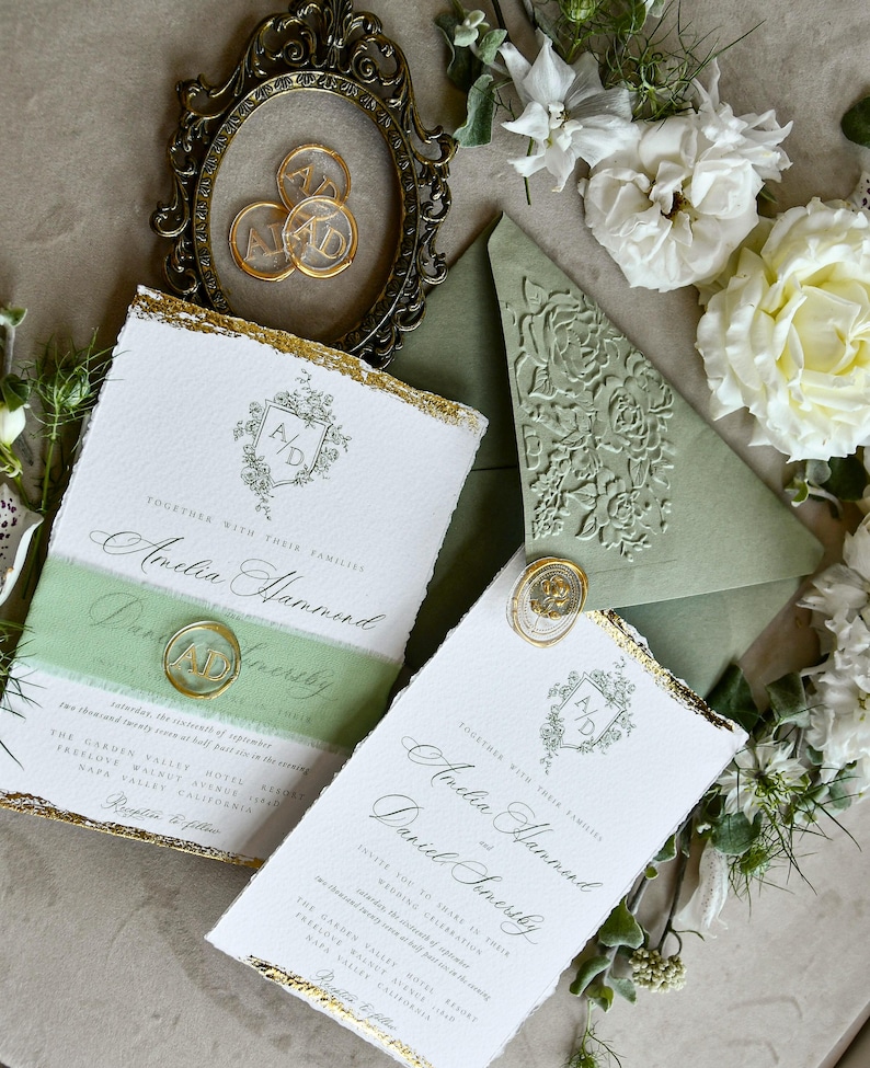 May include: Wedding invitation set featuring white textured paper with gold leaf edges, sage green belly bands, and wax seals with the initials AD. Includes a sage green envelope with a floral embossed flap and a vintage-style oval frame.