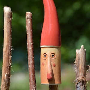 May include: A ceramic gnome figurine with a red hat and a brown face. The gnome is standing on a wooden stick.