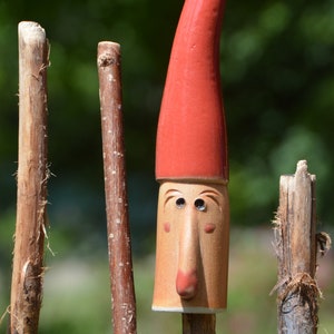 May include: A red ceramic gnome head with a long, pointed hat. The gnome has a large nose and small eyes. The gnome head is mounted on a wooden stick.