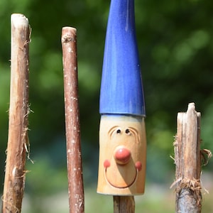 Puede incluir: Una figura de gnomo de cerámica con un sombrero azul y una cara sonriente. El gnomo está de pie sobre un palo de madera.