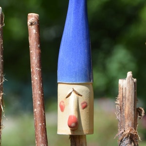 May include: A ceramic gnome figurine with a blue hat and a white face. The gnome is standing on a brown stick.