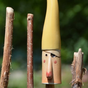 May include: A ceramic figurine of a person with a yellow hat and a brown face. The figurine has a black eye patch and a large nose. The figurine is attached to a brown stick.