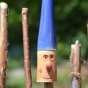 May include: A ceramic gnome figurine with a blue hat and a smiling face. The gnome is standing on a wooden stick.