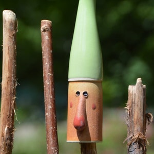 May include: A whimsical ceramic gnome figurine with a green hat and a brown face. The gnome is standing on a wooden stick.