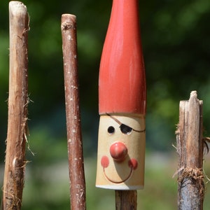 May include: A red ceramic gnome with a white face, black eye patch, and a red nose. The gnome is attached to a wooden stick.