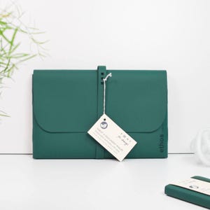 May include: A dark green tablet and document wallet with a flap closure and a strap. A white tag is attached with the text "VENT for change" and "TABLET & DOCUMENT WALLET". The wallet is made of a sustainable material.