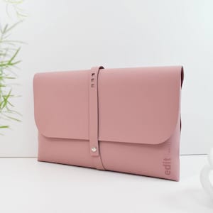 May include: A dusky pink portfolio case with a flap closure and a strap secured by a silver button. The strap has three small, evenly spaced cutouts. The word "edit" is printed vertically on the lower right corner.