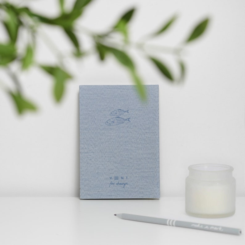 Puede incluir: Un cuaderno de lino azul con un dise&ntilde;o de dos peces y el texto "VENT for change".