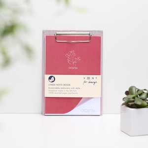 Peut inclure: Un carnet à lignes rose avec un clip argenté, un motif de feuille blanche et le texte "OCEAN" et "VNT for change".