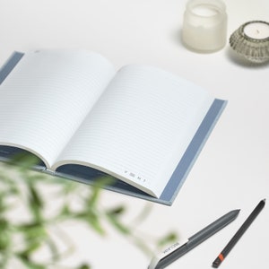 Puede incluir: Un cuaderno azul abierto con p&aacute;ginas rayadas y la palabra "VENT" impresa en la esquina inferior derecha. Dos bol&iacute;grafos est&aacute;n sobre una superficie blanca junto al cuaderno.