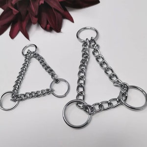 Könnte beinhalten: Zwei silberfarbene Ketten-Schlüsselanhänger. Jeder Schlüsselanhänger hat ein dreieckiges Kettendesign mit drei kreisförmigen Ringen. Die Ketten bestehen aus kleinen, ineinandergreifenden Gliedern. Die Schlüsselanhänger werden vor einem weißen Hintergrund mit einem dunkelroten floralen Akzent präsentiert.