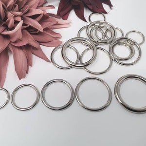 Könnte beinhalten: Eine Sammlung von silbernen Metall-Sprühringen in verschiedenen Größen. Die Ringe sind in einem zufälligen Muster auf einer weißen Oberfläche angeordnet.