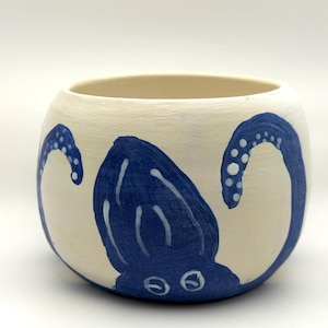 May include: A round, off-white ceramic bowl featuring a hand-painted blue octopus design. The octopus has a central body with two visible eyes and two curved tentacles extending upwards on either side, decorated with small white circular suction cups.