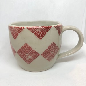 May include: A white ceramic mug with a red diamond pattern. The mug has a rounded shape and a handle.