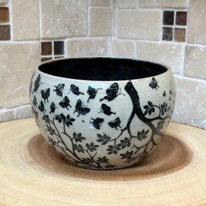 May include: A white ceramic bowl with a black and white design of butterflies and branches. The bowl is sitting on a wooden surface.