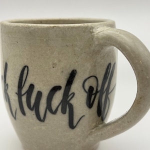 May include: A beige ceramic mug with a curved handle. The mug features the words "fuck off" in black cursive lettering. The mug has a speckled texture and a simple, functional design.