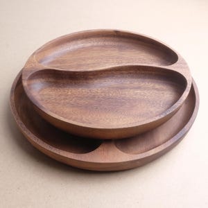 Puede incluir: Dos fuentes de madera, una apilada sobre la otra. La fuente superior tiene un divisor curvo, creando dos secciones. La veta de la madera es visible y el color es marrón cálido.