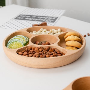 Puede incluir: Una bandeja redonda de madera para servir con cinco compartimentos separados. La bandeja está llena de varios bocadillos, incluyendo pistachos, piñones, galletas y rodajas de lima.