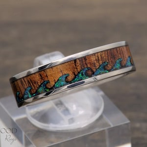 Puede incluir: Un anillo de plata con una banda de madera y un diseño de olas azul y verde. El anillo está en un expositor transparente.