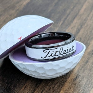 May include: A black and white ring with the word "Titleist" engraved on the white band. The ring is displayed in a golf ball ring box.