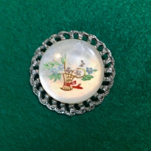 Puede incluir: Un broche redondo de tono plateado con un centro de nácar blanco que presenta un diseño floral pintado a mano. Las flores están en una cesta con un lazo rojo.