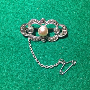 Puede incluir: Un broche de plata con un diseño delicado que presenta una perla falsa y una cadena con un pasador de seguridad.