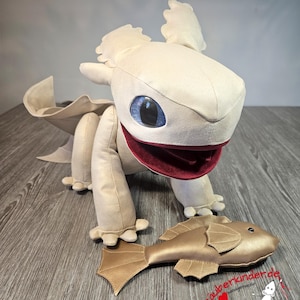 Op de afbeelding: Witte pluche draak met blauwe ogen en een rode mond, die een gouden pluche vis in zijn bek houdt. De draak zit op een houten oppervlak.