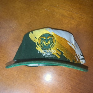 May include: Green and gold baseball cap with a Colorado State University logo and a ram mascot. The cap has a white and gold paint splatter design.