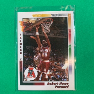 Op de afbeelding: Een vintage basketbal trading card met Robert Horry, een forward voor de Alabama Crimson Tide. De kaart is rood, wit en blauw met een foto van Horry in actie tijdens een wedstrijd. De kaart bevat de tekst "Wild Card" en "In Pride!"