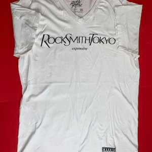 Op de afbeelding: Wit T-shirt met V-hals met de tekst "RockSmithTokyo" en "expensive" gedrukt op de voorkant. Het shirt heeft ook een zwart-witte "Parental Advisory Explicit Content" sticker rechtsonder.