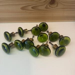 May include: A collection of ten round cabinet knobs featuring translucent olive green glass tops and brass-toned metal bases. Each knob has a smooth, circular surface and a short metal screw extending from the back for mounting purposes.