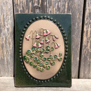 May include: A square ceramic tile with a botanical design. The tile features a central oval with a beige background, showcasing pink bell-shaped flowers and green leaves. The oval is framed by a dark green border with a dotted pattern.