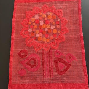 May include: A red textile with a floral design. The flower has a circular head with a pattern of orange, pink, and red squares. The stem and base are also red. The edges are finished with a red border.