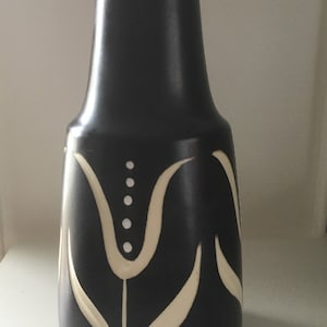 May include: A tall, dark vase with a flared base and a narrow neck. The vase is black with a cream-colored floral design. The design includes stylized tulip-like shapes and a series of dots. The vase is likely ceramic.