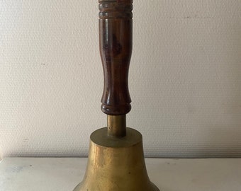 May include: A brass bell with a wooden handle. The bell is gold in color and has a rounded shape. The handle is brown and has a knob on top.