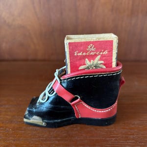 May include: A black leather boot with red accents holds a matchbox with the text "Solo Edelweiss" and a picture of a white flower.