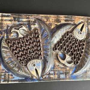May include: Rectangular ceramic wall art depicting two stylized fish. The fish have textured, brown scales and light blue and cream details. The background features a blue and brown geometric pattern. The art is approximately 25 cm wide.