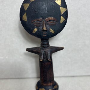 May include: A wooden figurine of a person with a round head and a stylized face. The head is decorated with gold triangles. The figurine is standing with its arms raised and its hands clasped together. The figurine is painted black with gold accents.