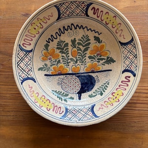 Può includere: Piatto decorativo in ceramica con un motivo floreale. Il piatto presenta una base color crema con un bordo blu e rosso a motivi. Il centro del piatto mostra un bouquet di fiori arancioni e foglie verdi.