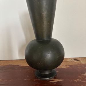 Könnte beinhalten: Eine dunkelbronze Vase mit kugelförmigem Sockel und konischem Oberteil. Die Vase hat eine dunkle, gealterte Patina und steht auf einem kleinen, kreisförmigen Sockel. Die Gesamtform ist eine einzigartige Kombination geometrischer Formen.