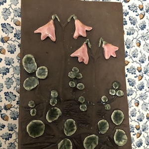 May include: A rectangular, brown ceramic tile with raised floral and leaf designs. Pink bell-shaped flowers with green stems and leaves are arranged across the surface. The tile has a glossy finish and is likely handmade.