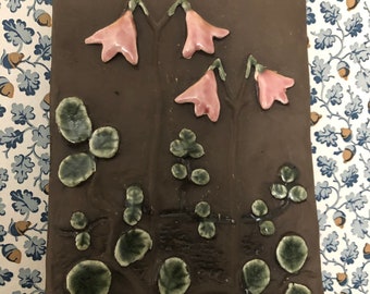 May include: A rectangular, brown ceramic tile with raised floral and leaf designs. Pink bell-shaped flowers with green stems and leaves are arranged across the surface. The tile has a glossy finish and is likely handmade.